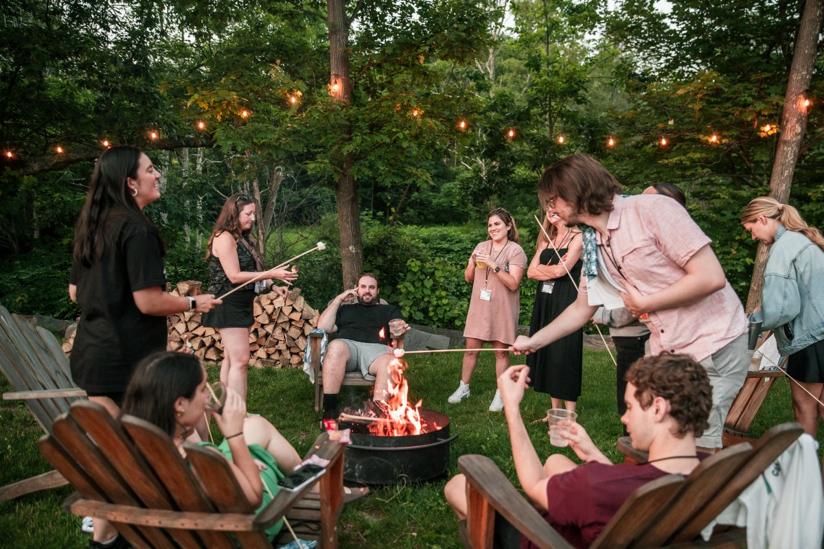 A group of people sit around a campfire, they are boosting employee morale by making s'mores.